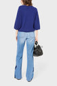 Person standing facing away, wearing a blue sweater and light blue wide-leg jeans with dark patches on the sides, holding a black handbag in their right hand. The background is plain white.