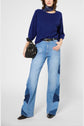 Person wearing a navy blue sweater, light blue wide-leg jeans with dark blue patches, black pointed shoes, a neck scarf, and holding a black handbag. The background is plain and light.