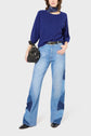 Woman wearing a blue sweater, patchwork light blue jeans, black pointed shoes, a dark scarf around her neck, and holding a black handbag. She stands against a plain white background.