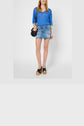 Woman wearing a blue long-sleeve top, denim mini skirt, black wedge sandals, and carrying a black shoulder bag, standing against a plain white background.