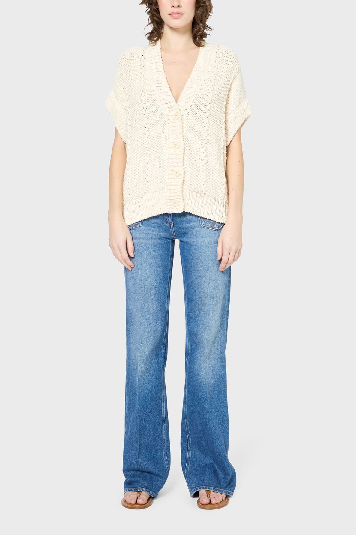 A person wearing a cream short-sleeve knit cardigan, blue flared jeans, and brown sandals stands against a plain light grey background.