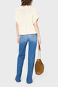 A person with shoulder-length hair, wearing a cream knit top and blue jeans, stands facing away while holding a brown suede bag with a chain strap.
