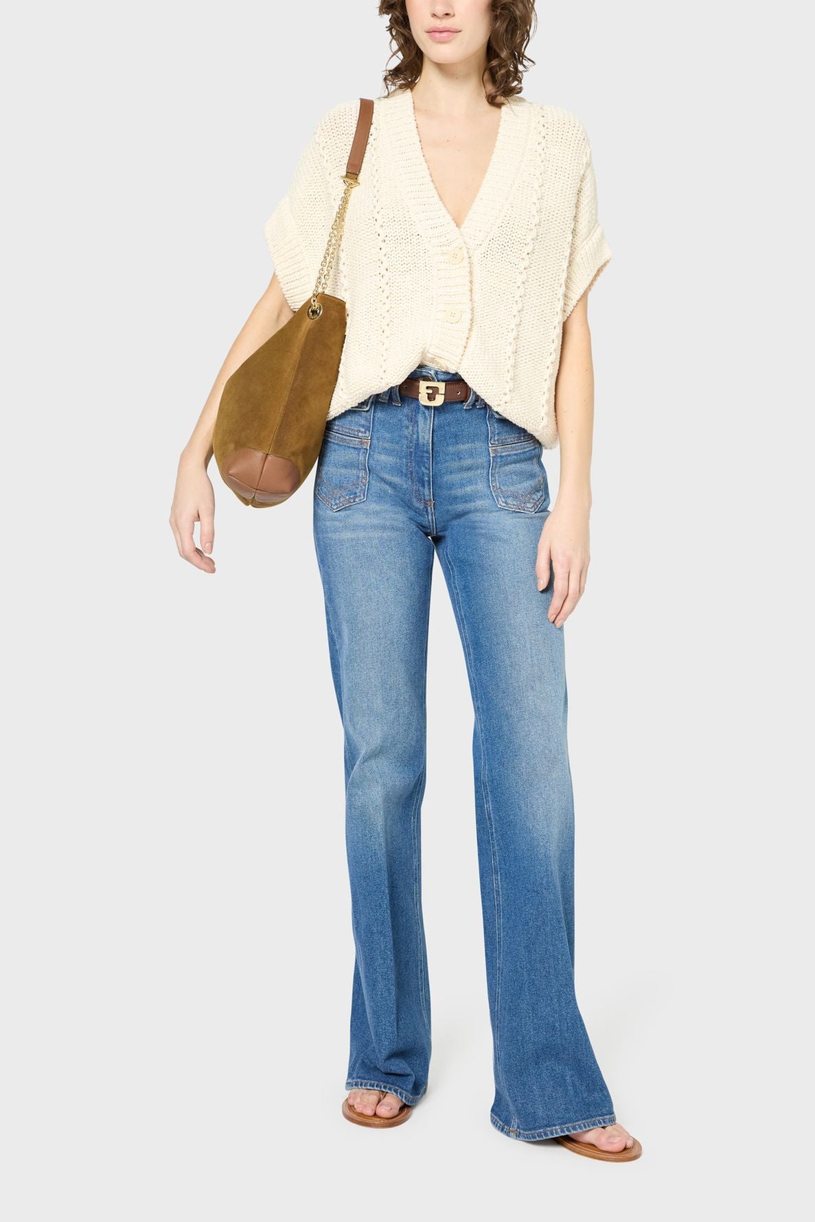 A person wearing a cream knit short-sleeve sweater, blue flared jeans, brown belt, brown shoulder bag, and sandals stands against a plain background.