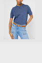 Person wearing a short-sleeve blue knit shirt, light blue jeans with large front pockets and frayed edges, and a tan belt with gold hardware, posing with one hand on hip against a plain background.