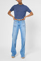 Person wearing a short-sleeved blue top, light blue wide-leg jeans with visible front pockets and white side seams, brown belt, and beige shoes, standing with hands on hips against a plain background.