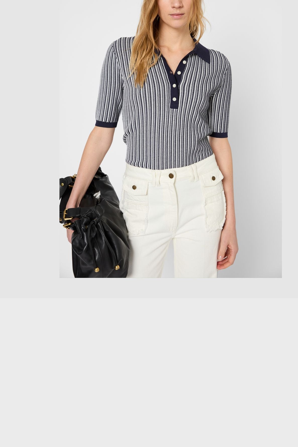Woman in a short-sleeved, navy and white striped polo shirt and high-waisted white pants with front pockets, holding a black handbag. She stands against a plain light background.