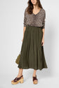 A woman wearing a col V pull léopard, an olive green pleated skirt, and brown platform sandals stands against a neutral background, holding a small textured bag at her side.
