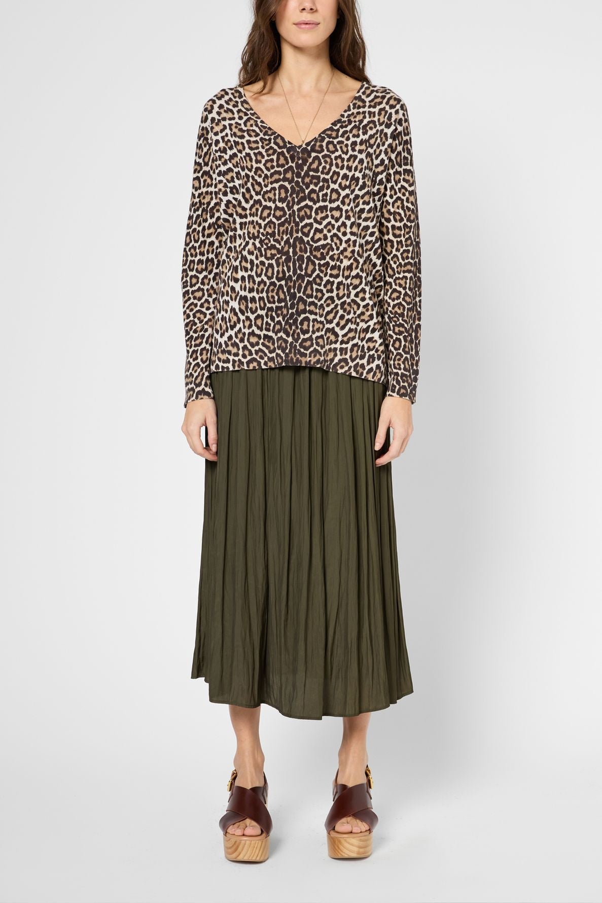 A woman wears a col V pull léopard à manches longues, an olive green pleated midi skirt, and brown platform sandals. She stands against a plain light background.