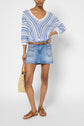 A person wearing a blue and white pull rayé col V sweater, denim mini skirt, brown sandals, and holding a woven bag stands against a plain background.