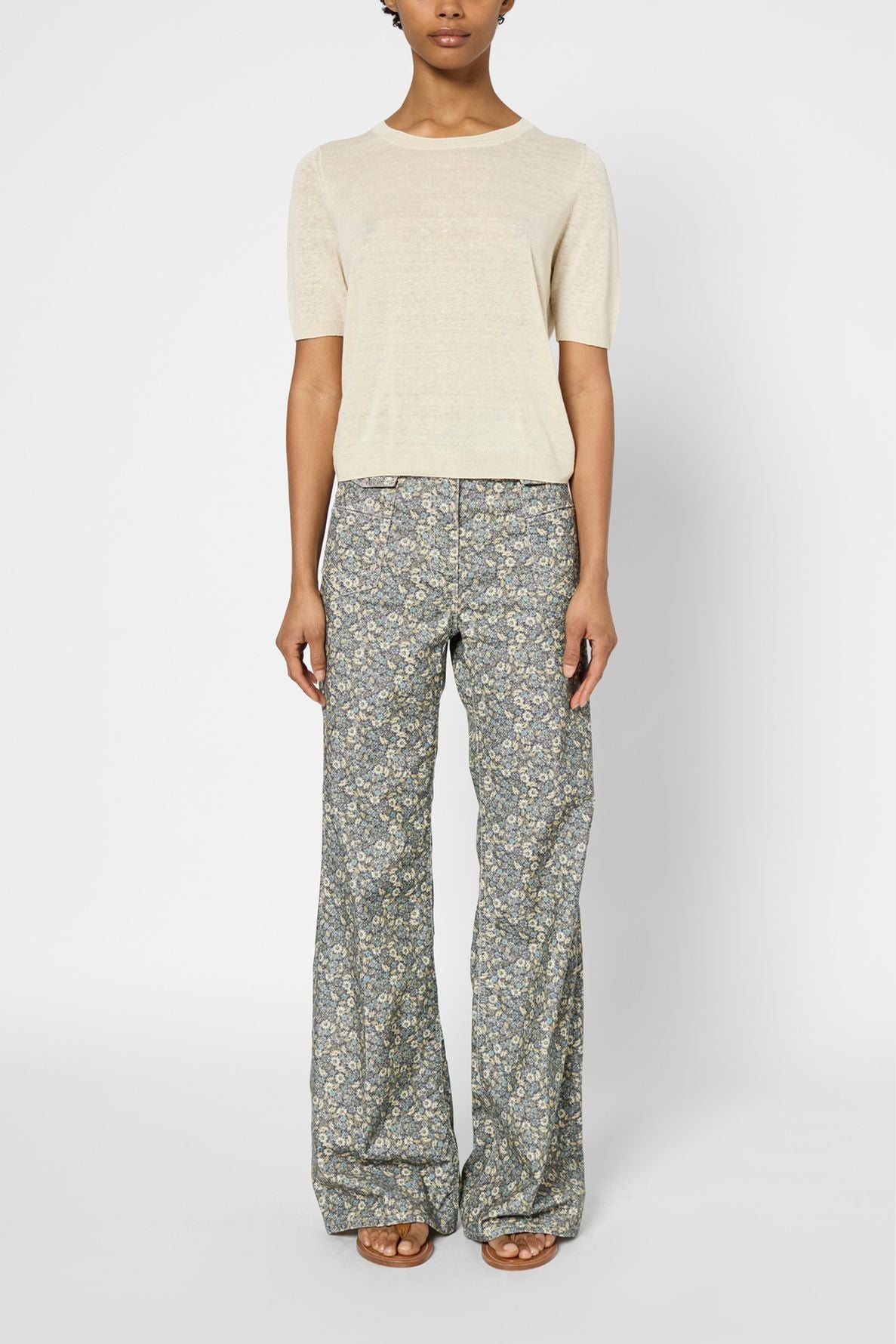 A person stands facing forward, wearing a pull manches courtes beige top and high-waisted, wide-leg floral pants with sandals. The background is plain white.