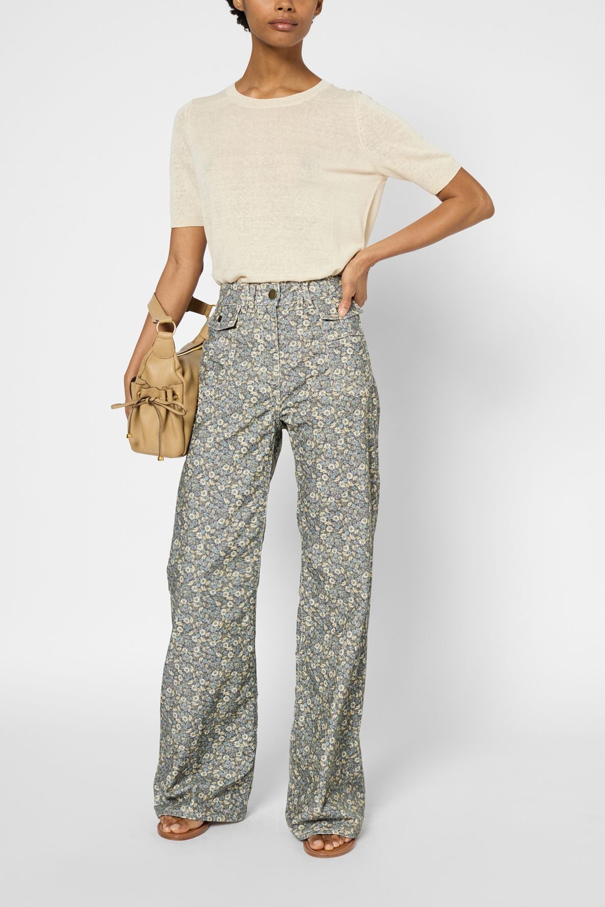 Woman wearing a sheer cream t-shirt reminiscent of a pull manches courtes, high-waisted floral wide-leg pants, sandals, and holding a tan handbag stands against a plain background.