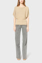 A person stands facing forward, wearing a beige textured short-sleeve sweater with a subtle col bateau neckline and gray patterned wide-leg pants featuring a geometric print. The background is plain light gray. This elegant pull maille dorée is soigneusement fabriqué en Italie.
