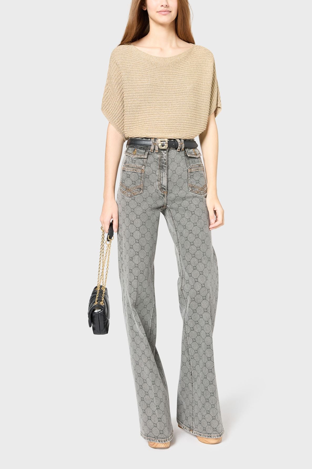 A woman wearing a beige col bateau knit top, high-waisted patterned gray flared jeans, and holding a black purse with a gold chain strap stands against a plain light background.