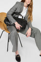 A woman in a grey blazer à imprimé caviar sits on a chair, holding a large black handbag. She has long, light brown hair and wears black loafers. The background is plain and white.
