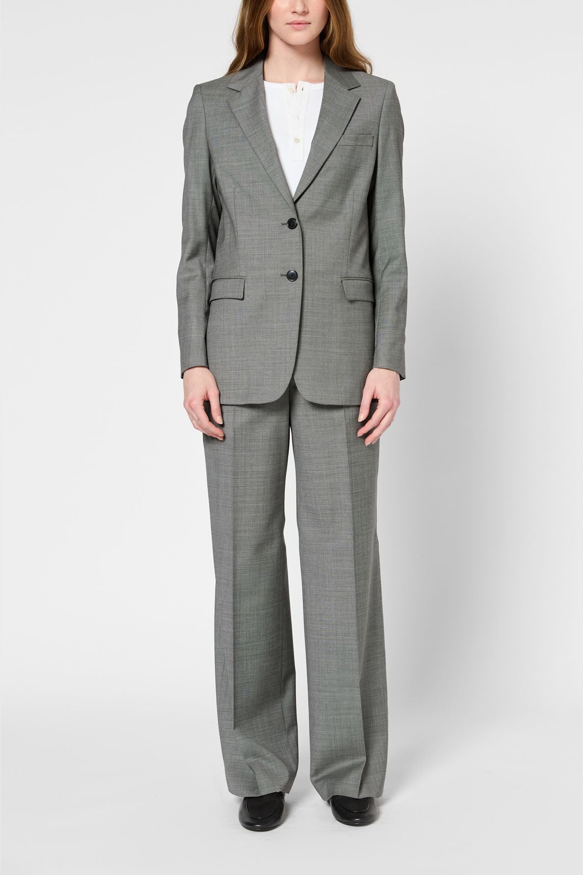 A person stands facing forward, wearing a gray blazer femme with wide-leg trousers, a white shirt underneath, and black shoes, against a plain white background.