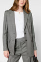 A person wearing a light gray veste tailleur and matching pants over a white buttoned shirt, holding a black bag with one hand, against a plain background. Only the upper body is visible.