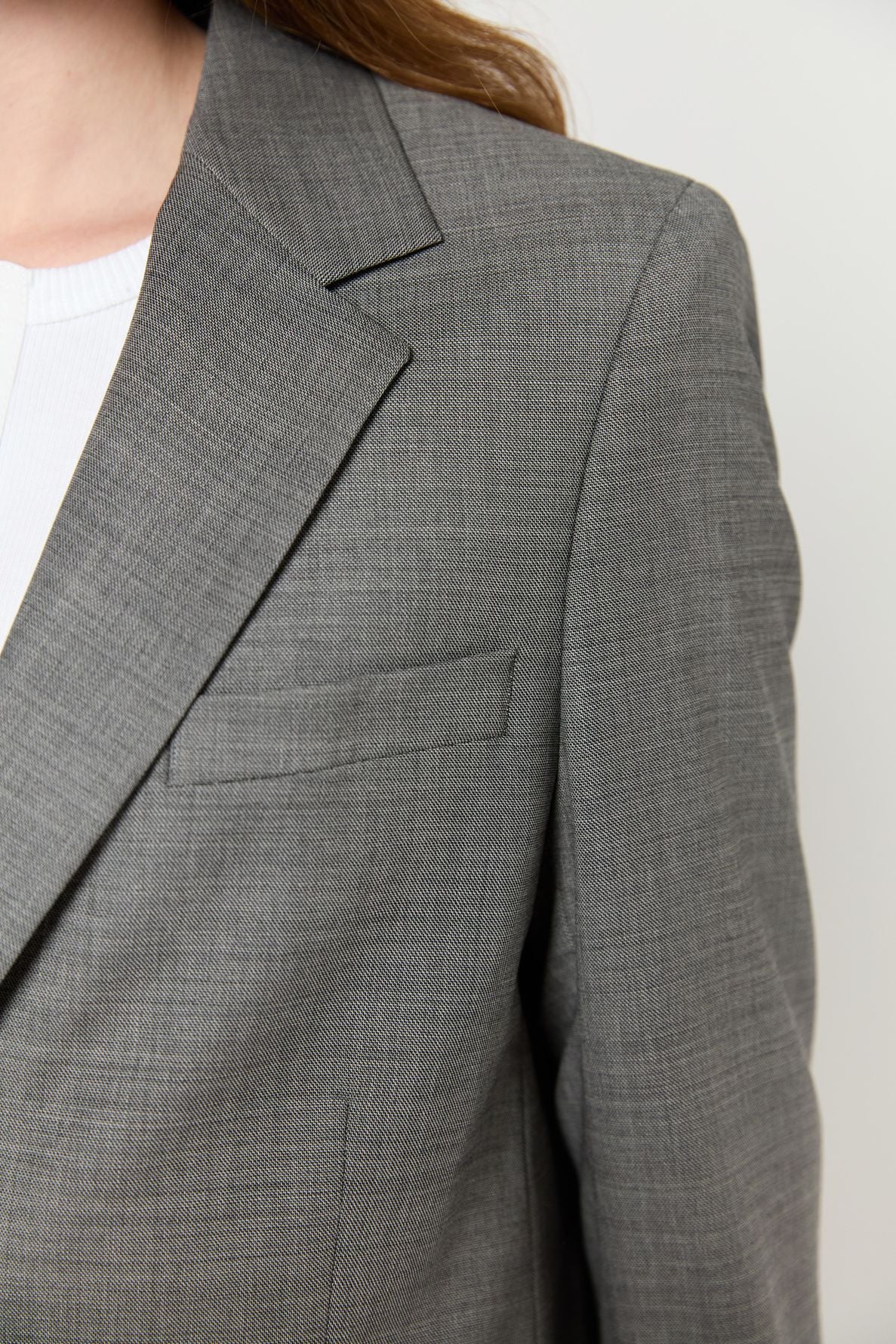 Close-up of a person wearing a gray blazer femme over a white top. Only part of the person’s chest, shoulder, and hair are visible, with a focus on the veste tailleur’s lapel and pocket.