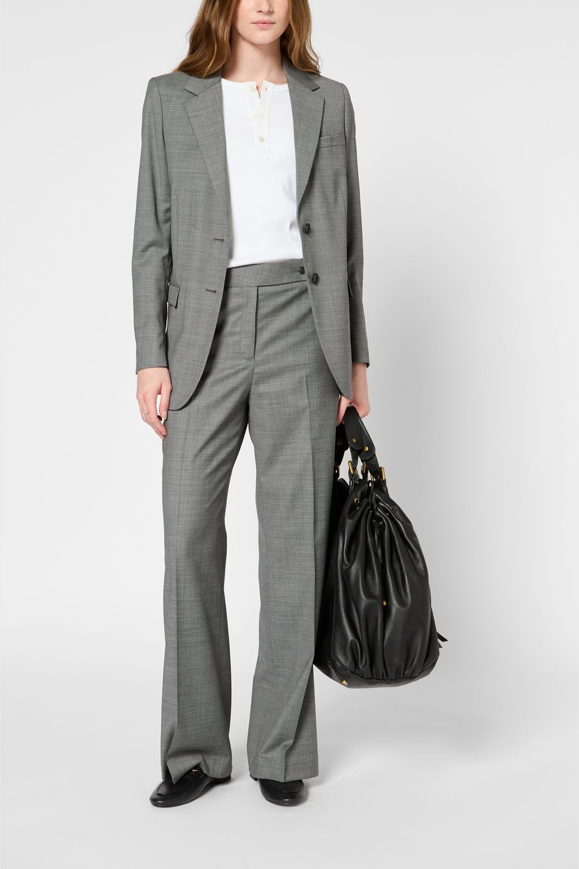 A woman in a gray blazer à imprimé caviar pantsuit and white blouse stands facing forward, holding a large black handbag in her left hand. She wears black shoes. The background is plain and white.