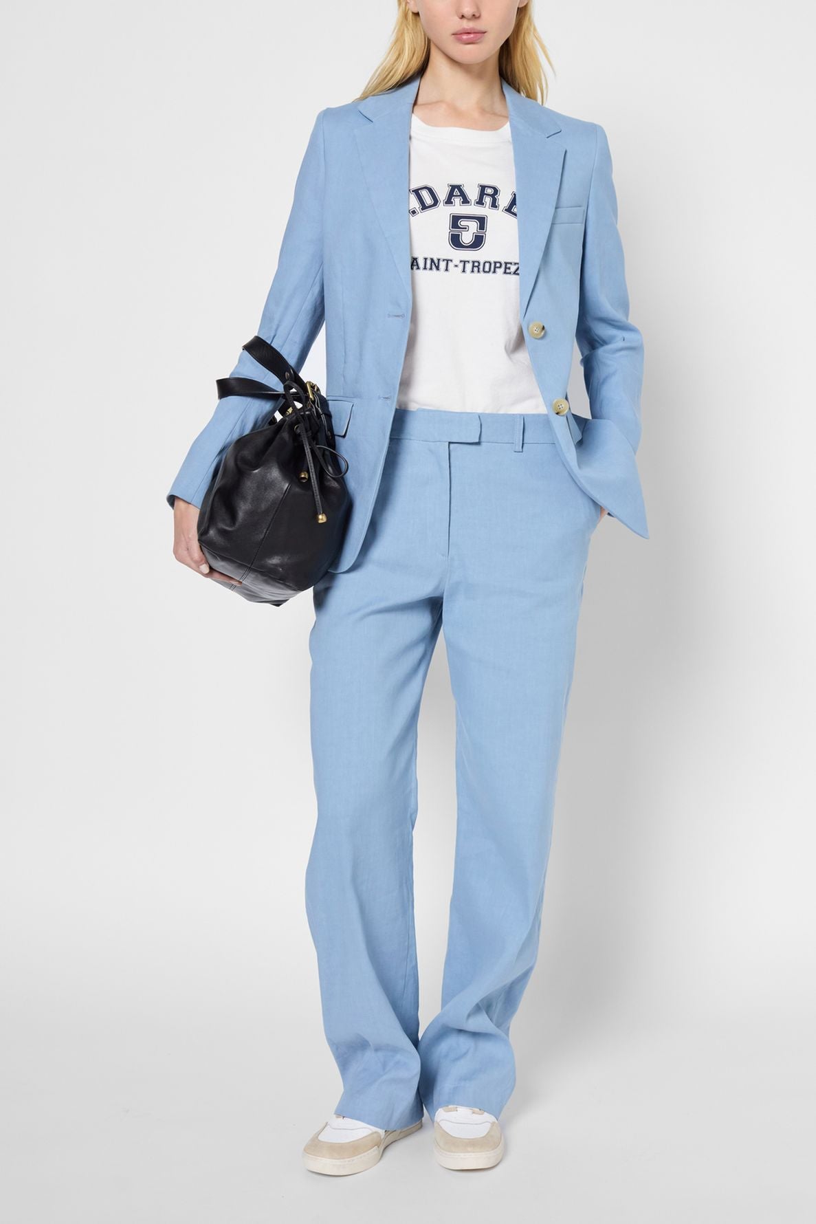 A person wearing a light blue blazer en lin, a white printed t-shirt, and white sneakers holds a black handbag. The background is plain and light-colored.