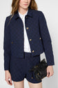 A person wearing a navy blue textured jacket and matching shorts over a white shirt holds a small black handbag.