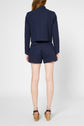 A person stands facing away, wearing a navy textured jacket and matching high-waisted shorts, paired with black platform sandals with wooden heels. The background is plain white.