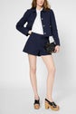 A woman wears a navy jacket and shorts set with a white shirt, holding a small black bag. She has light skin and brown wavy hair, and is wearing black platform sandals with wooden heels.