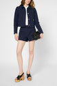 A woman wearing a navy patterned jacket and matching shorts, a white shirt, black wooden platform sandals, and holding a small black purse, poses against a plain light background.
