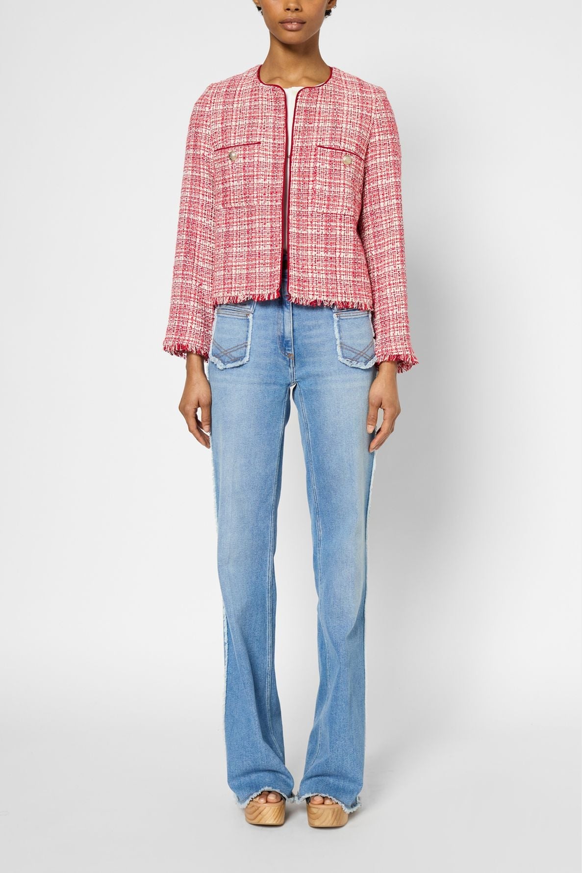 A person wearing a veste en tweed rose chic with front pockets, light blue flared jeans with patch pockets, and beige shoes, standing against a plain background.