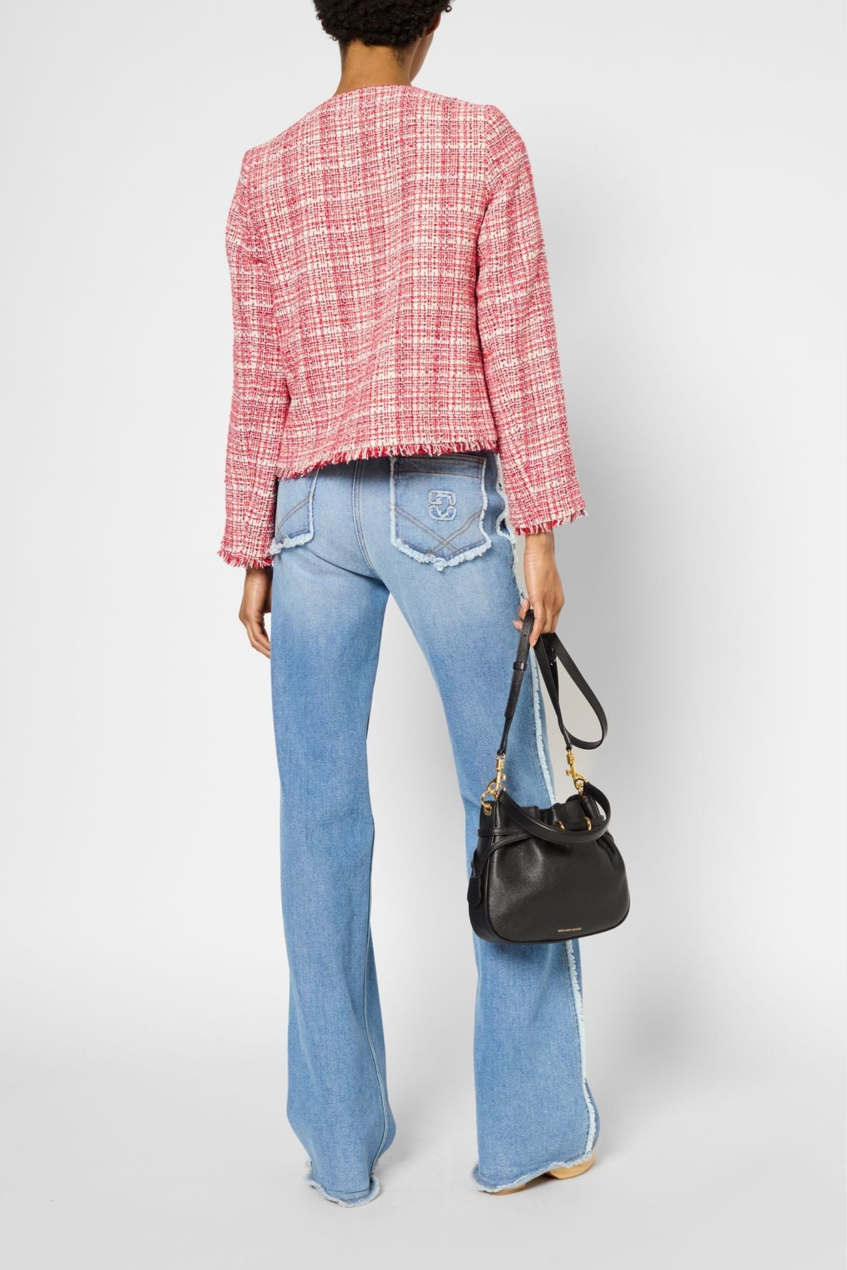 A person stands facing away, wearing a veste courte femme in pink tweed and light blue flared jeans, holding a black handbag in their left hand. The background is plain white.