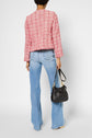 A person stands facing away, wearing a veste courte femme in pink tweed and light blue flared jeans, holding a black handbag in their left hand. The background is plain white.