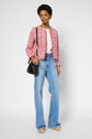 Woman wearing a chic veste en tweed rose, white top, light blue high-waisted jeans, and beige flats stands against a plain background, holding a black shoulder bag.