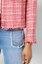 A person wears a veste courte femme—a chic red and white tweed jacket with a frayed hem—paired stylishly with light blue distressed jeans. Only the lower torso, arm, and hand are visible against a plain background.
