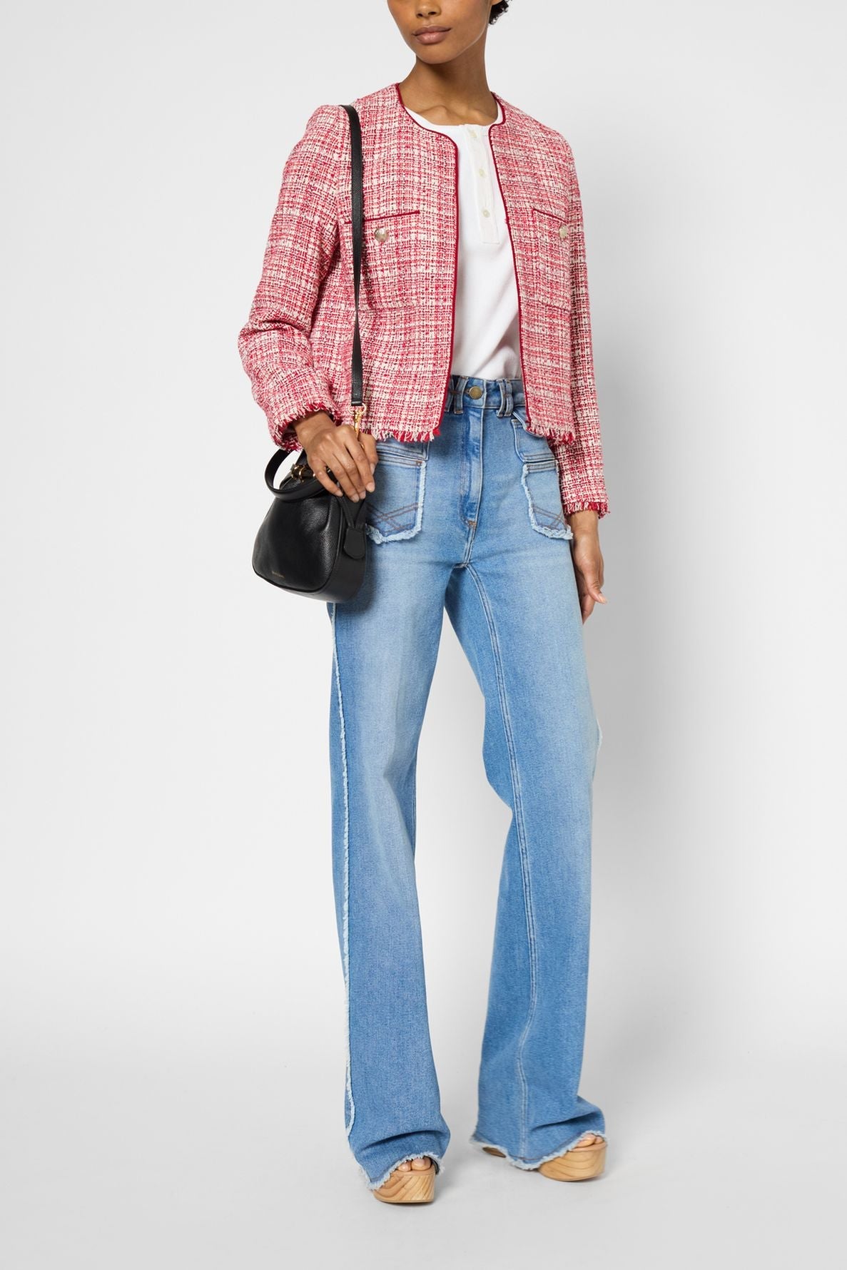 A person stands wearing a pink veste en tweed courte femme, white shirt, light blue high-waisted jeans with large front pockets, beige shoes, and carries a small black handbag.