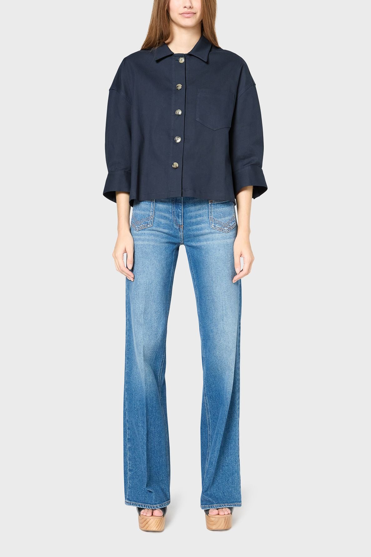 Woman wearing a navy button-up shirt with wide sleeves, styled like a surchemise coton, and light blue high-waisted flared jeans, standing against a plain light gray background. Her face is partially out of frame.
