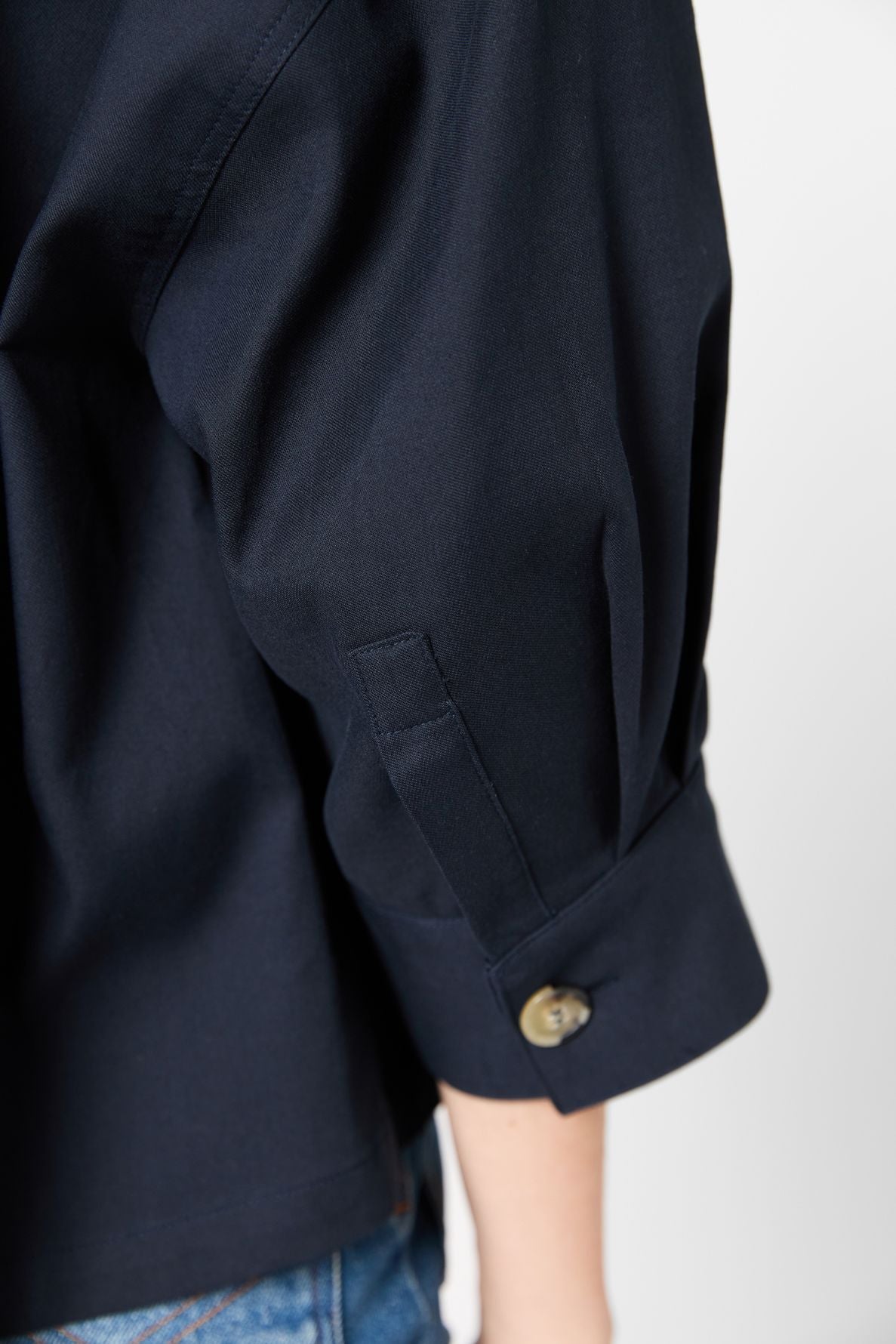 Close-up of a person wearing a navy blue surchemise coton with cuff and button detail, paired with blue denim jeans. The shirt sleeve is slightly rolled up, exposing part of the forearm.