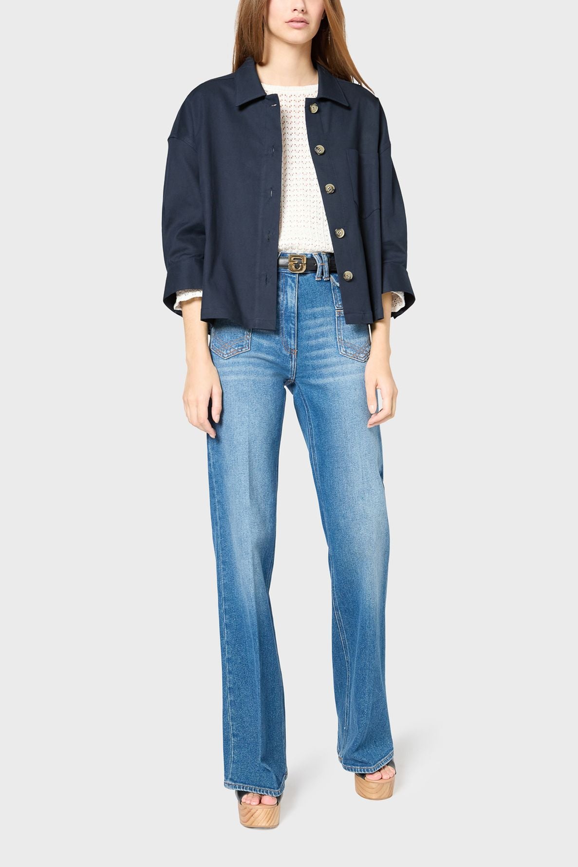 A woman stands wearing a veste courte navy jacket, white textured top, high-waisted blue jeans, and platform sandals against a plain light gray background.