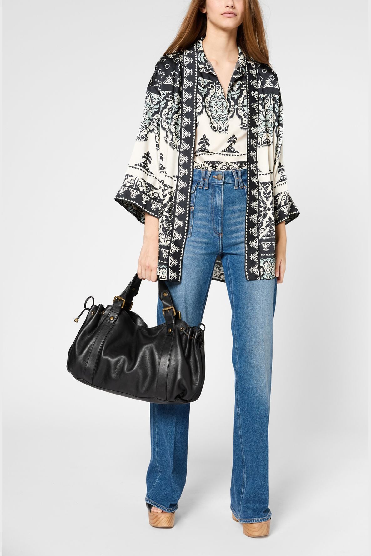 Woman wearing a patterned kimono-style top, high-waisted blue jeans, and tan platform shoes, holding a large black handbag against a plain background.