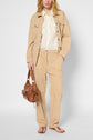 A person wearing a beige coton lyocell lin veste surchemise, a white shirt, beige pants, and flip-flops holds a brown handbag with gold accents. The background is plain and light-colored.