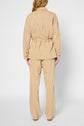 A person with long, wavy hair is standing facing away, wearing a loose-fitting beige veste surchemise with a cinched waist and matching beige pants crafted from a soft coton lyocell lin blend. The background is plain and white.