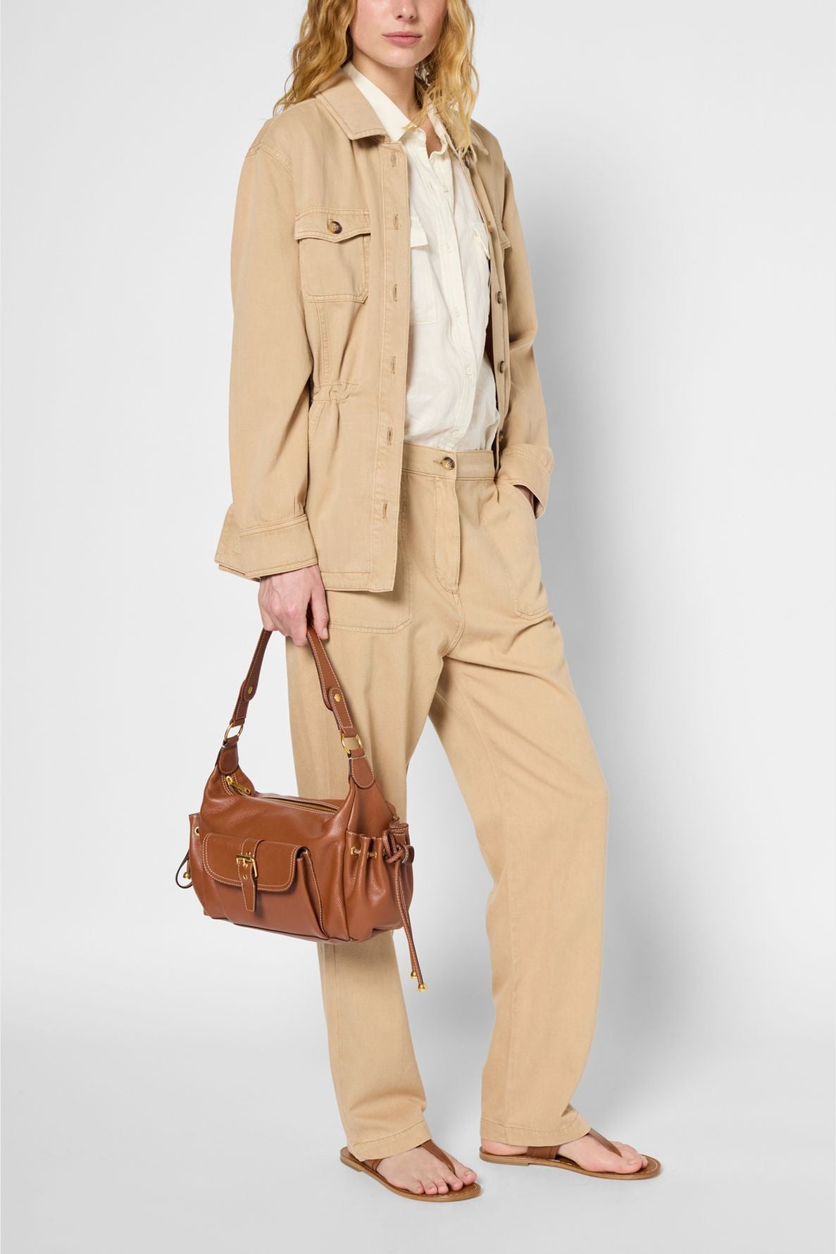 A person wearing a beige veste cintrée and pants, a white shirt, and sandals, holding a brown leather shoulder bag. The outfit has a casual, neutral-toned style that highlights the refined silhouette of the fitted jacket.