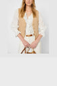 Woman wearing a beige gilet court en coton et lin over a white ruffled blouse and white pants, holding a brown-handled bag. Her hair is loose, and she is standing against a plain light background.