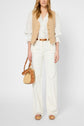 A woman wears a beige gilet sans manche over a white blouse, paired with white wide-leg pants and platform sandals. She carries a small brown and woven handbag. Her hair is loose, and her face is not visible.