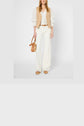 A person wearing a beige gilet court en coton et lin over a white blouse with puffed sleeves, white wide-leg pants, brown sandals, and holding a small tan handbag.