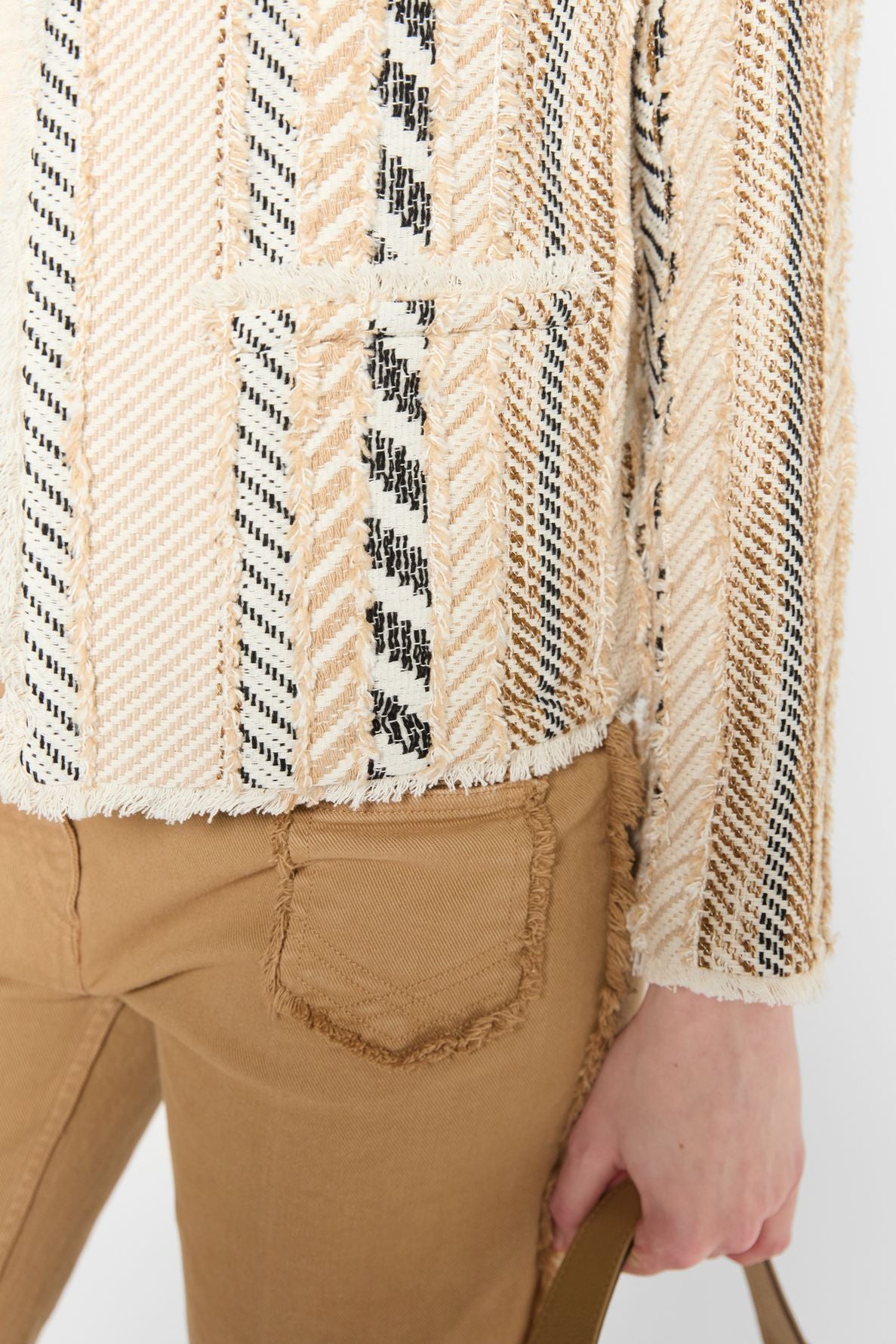 A person wearing camel-colored pants with a frayed pocket and a cream, tan, and black textured jacket with a striped pattern is holding a tan strap. The background is white.