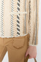 A person wearing camel-colored pants with a frayed pocket and a cream, tan, and black textured jacket with a striped pattern is holding a tan strap. The background is white.