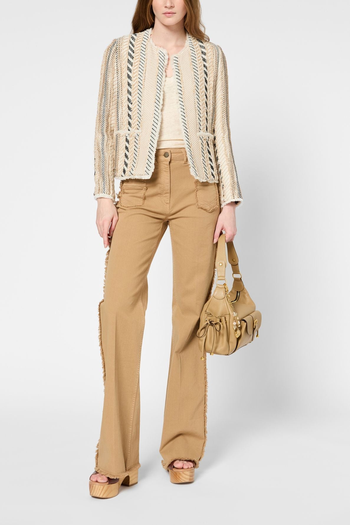 A person wearing a textured cream and beige jacket, a white top, tan wide-leg pants with frayed sides, beige platform sandals, and holding a tan handbag, standing against a plain background.