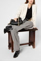 A person sitting on a wooden bench wears a white plaid blazer, black top, gray wide-leg pants, and black boots, holding a black handbag with a gold chain strap.