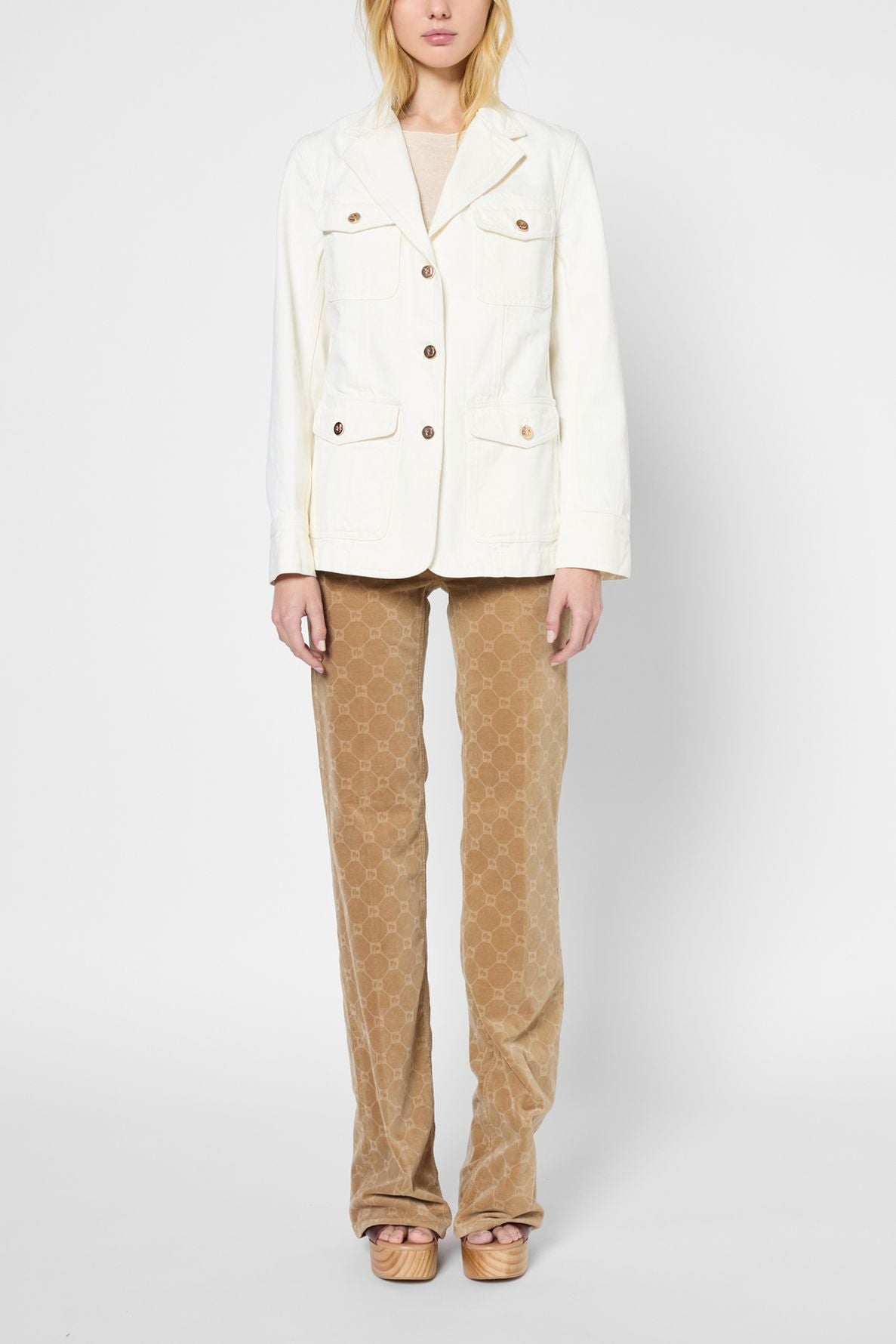 A woman stands facing forward, wearing a cream blazer, light top, tan patterned pants, and platform sandals against a plain white background. Her hands are relaxed at her sides.