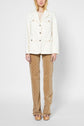 A woman stands facing forward, wearing a cream blazer, light top, tan patterned pants, and platform sandals against a plain white background. Her hands are relaxed at her sides.