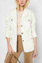 A woman wearing a cream blazer over a beige top and tan patterned pants holds a beige bag. Only the lower part of her face is visible.